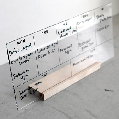 The Girl Friday Weekly Black Acrylic Calendar with Wood Stand displays handwritten tasks in black marker, listing activities like Drive Carpool, Rehearsal, and Date Night from Monday to Sunday. Its a reusable dry-erase planner set on a gray surface.