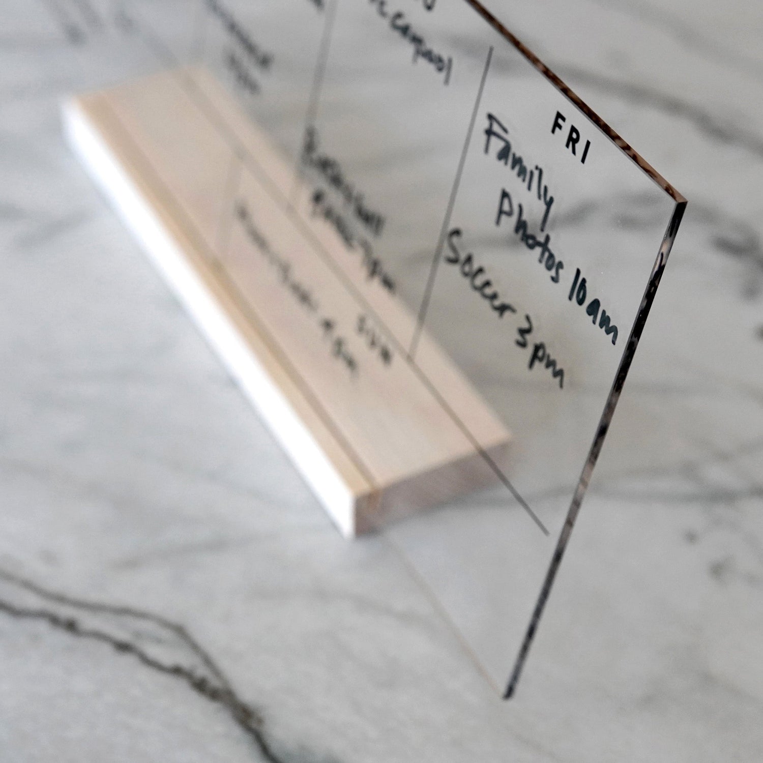 The Weekly Black Acrylic Calendar with Wood Stand by Girl Friday displays handwritten notes like FRI: Family Photos 10am, Soccer 3pm. It rests elegantly on a marble surface, stylishly organizing your week.