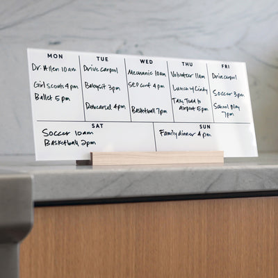 The Girl Friday Weekly White Acrylic Calendar with Wood Stand is a modern desk accessory that stands out on a marble surface. Perfect for organizing appointments and activities, it neatly lists events like Soccer at 10am and Family dinner at 4pm.