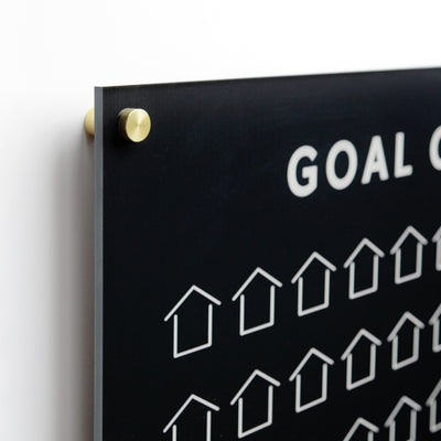 A close-up of the Goal Chart | Sales Tracker | Black Acrylic by Girl Friday shows a black board with the word GOAL among house icons, elegantly mounted on a wall with gold-colored pegs.