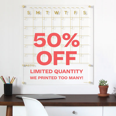 A 26 x 30 Clear Acrylic Monthly Wall Calendar by Girl Friday Shop, featuring gold text and days of the week above an empty grid. 50% OFF, Limited Quantity, and We printed too many! are in red, while a desk with pencils, wet-erase markers, and a small plant is beneath.