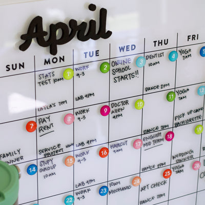 The Girl Friday Shops White Acrylic Monthly Calendar for April offers a sleek dry erase surface for vibrant markers and magnets or command strip mounting to securely display packed schedules, like tests, work shifts, online school starts, appointments, and dance sessions.