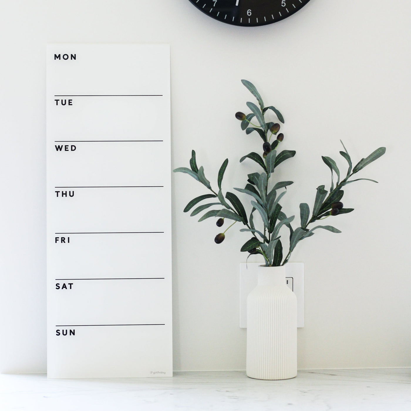 The White Acrylic Dry Erase Weekly Calendar from Girl Friday Shop showcases black text in a vertical Monday-to-Sunday format on a sleek white acrylic. A nearby white vase holds green leaves and berries, while a black clock peeks above, ideal for those preferring a stylish no-mount planner.