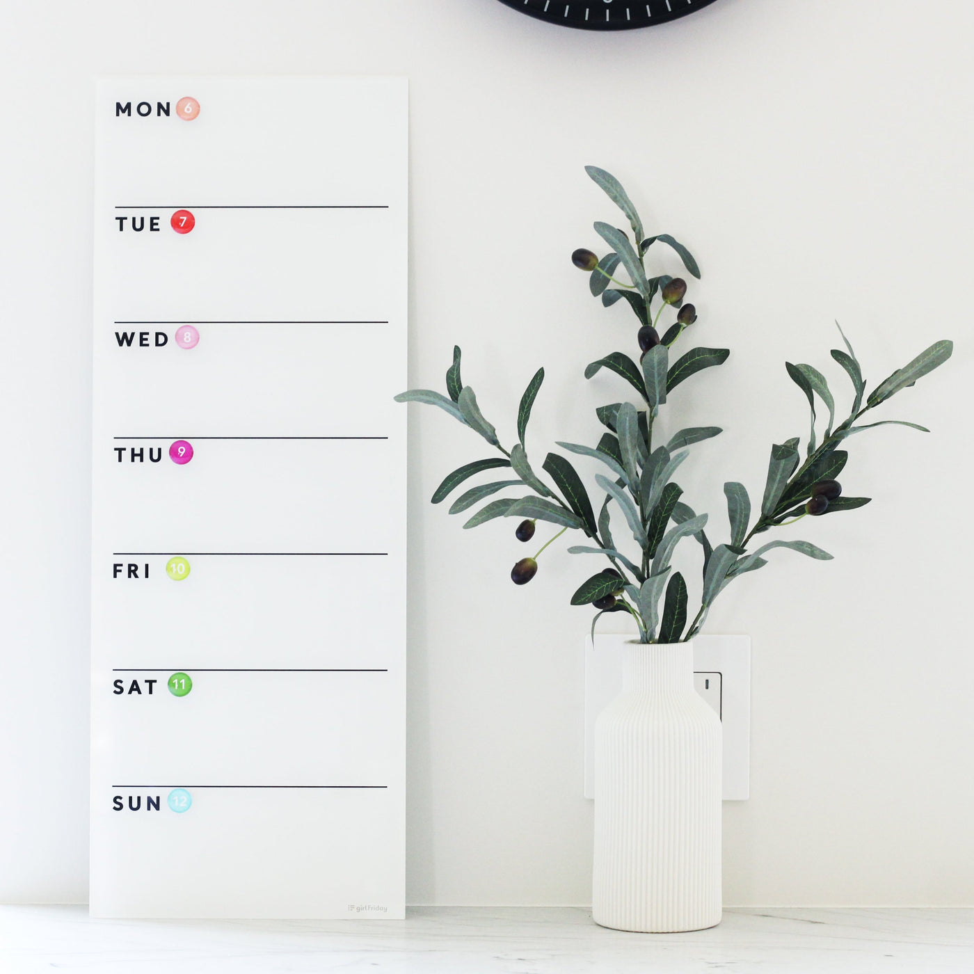The White Acrylic Dry Erase Weekly Calendar by Girl Friday Shop is chic and unmarked, hanging elegantly on a white wall next to a vase of olive branches. With vertically aligned days and colorful dots, it creates a stylish and organized space without the need for mounting.