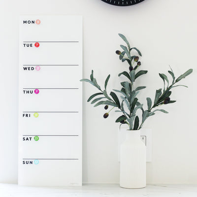 The White Acrylic Dry Erase Weekly Calendar by Girl Friday Shop is chic and unmarked, hanging elegantly on a white wall next to a vase of olive branches. With vertically aligned days and colorful dots, it creates a stylish and organized space without the need for mounting.