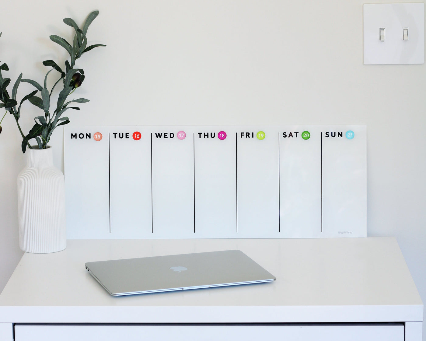 A closed laptop rests on a white desk, with a Girl Friday Shops White Acrylic Dry Erase Weekly Calendar positioned elegantly behind it. To the left is a white vase with green foliage. The pristine white wall features a convenient light switch on the right.