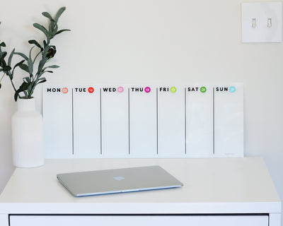 A closed laptop rests on a white desk, with a Girl Friday Shops White Acrylic Dry Erase Weekly Calendar positioned elegantly behind it. To the left is a white vase with green foliage. The pristine white wall features a convenient light switch on the right.
