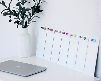 On a white desk, an open laptop sits beside a Girl Friday Shops White Acrylic Dry Erase Weekly Calendar. The mount-free planner spans Monday to Sunday. Next to it is a white vase with greenery, enhancing organization with its easy-to-update surface.