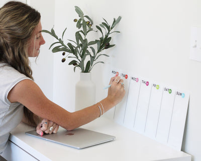 A woman uses the Girl Friday Shops White Acrylic Dry Erase Weekly Calendar, mounted horizontally, to organize her schedule. Days of the week are colorfully displayed as she enjoys wearing casual clothing and jewelry. Nearby, a vase with olive branches decorates her desk beside a closed laptop.