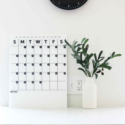 On a countertop sits the Girl Friday Shops White Acrylic Monthly Calendar, featuring blank spaces for planning. Beside it is a ribbed vase with leafy branches. Above, part of a wall clock is visible, and a nearby power outlet adds convenience.