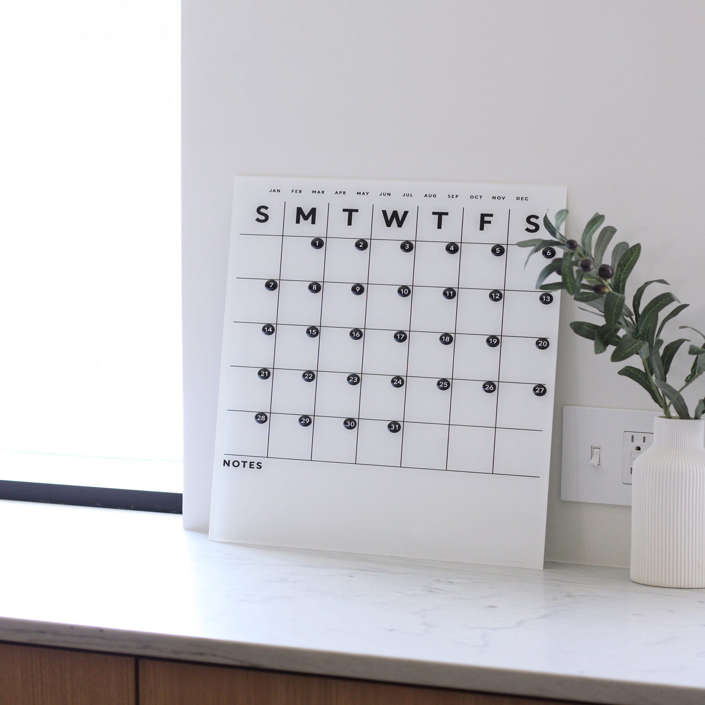 The Girl Friday Shops White Acrylic Monthly Calendar with Bottom Notes elegantly rests on a marble counter, featuring days marked from Sunday to Saturday in black letters. Beside it, a white vase with greenery adds natures touch, while a power outlet is discreetly positioned behind the display.