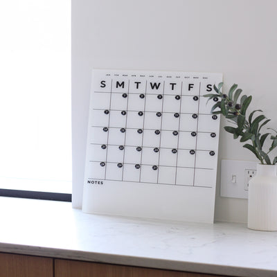 The Girl Friday Shops White Acrylic Monthly Calendar with Bottom Notes elegantly rests on a marble counter, featuring days marked from Sunday to Saturday in black letters. Beside it, a white vase with greenery adds natures touch, while a power outlet is discreetly positioned behind the display.