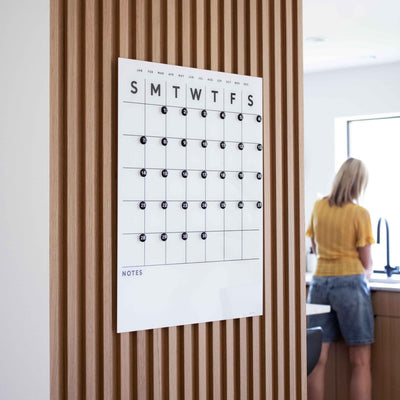 The Girl Friday Shops White Acrylic Monthly Calendar features bottom notes and mounts seamlessly on wood, with integrated whiteboard and magnets. In the bright room, a person in denim shorts and a yellow shirt stands at the sink.