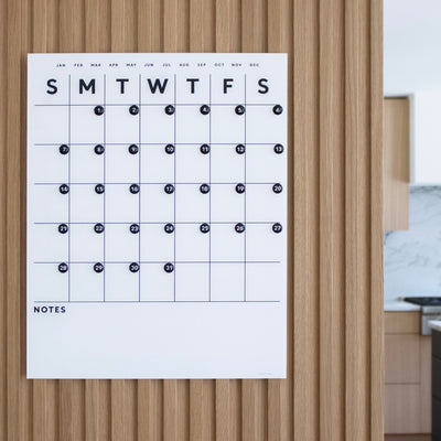 The White Acrylic Dry Erase Monthly Calendar by Girl Friday Shop is stylishly mounted with Command Strips in a kitchen. Featuring a wooden-backed surface, it displays day columns, month rows, black markers on dates, and a Notes section at the bottom.