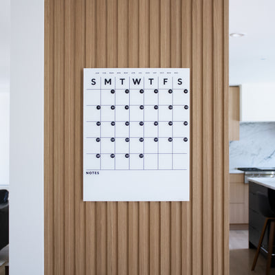 The White Acrylic Monthly Calendar from Girl Friday Shop, featuring days of the week across the top and black dots for dates, is mounted on a vertical wooden panel. In the background, a modern kitchen with light wood cabinetry can be seen.