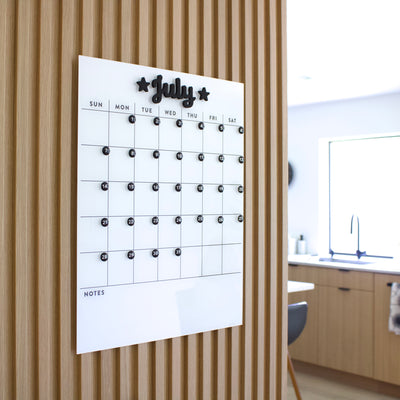 The White Acrylic Dry Erase Monthly Calendar by Girl Friday Shop is wall-mounted for July on a wooden panel. It features a dry erase grid, circular magnets for dates, space for notes, and is secured with command strips in a modern kitchen with light wood cabinets in the background.