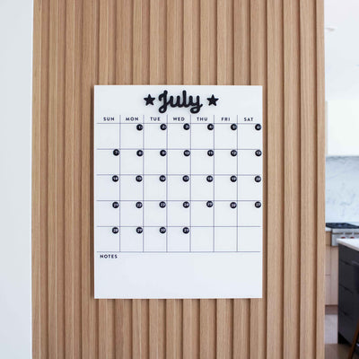 A White Acrylic Monthly Calendar by Girl Friday Shop, featuring a grid layout and month words with star accents, hangs on a light wooden wall. Each day includes space for notes with black markers. Its mounted using command strips, partially revealing the modern kitchen on the right.
