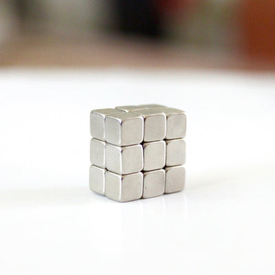 A small cube of 27 silver metallic pieces sits at the center of a white surface, resembling the intricate design of Girl Fridays Ultra Strong Neodymium Rare Earth Magnets, set against a softly blurred background.