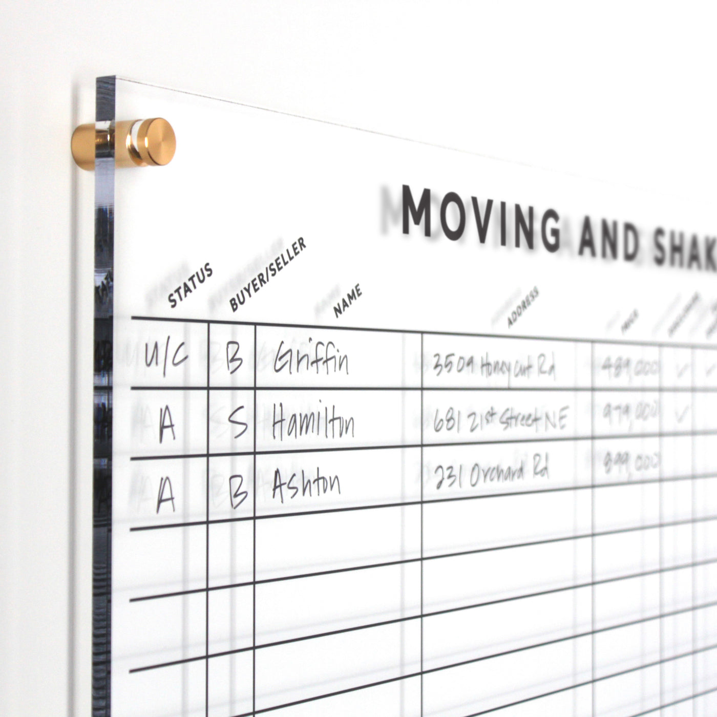 A close-up of the Moving & Shaking Clear Acrylic Real Estate Board by Girl Friday shows columns for Status, Buyer/Seller, Name, and Address with names like Griffin and Hamilton. The board includes gold standoffs for better sales tracking.