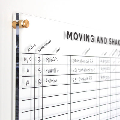 A close-up of the Moving & Shaking Clear Acrylic Real Estate Board by Girl Friday shows columns for Status, Buyer/Seller, Name, and Address with names like Griffin and Hamilton. The board includes gold standoffs for better sales tracking.