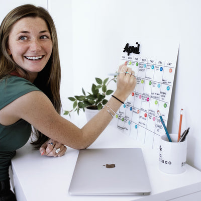 A person with long hair smiles while jotting notes on a White Acrylic Monthly Calendar by Girl Friday Shop for April. At a white desk, they’re accompanied by a closed laptop, pen-filled cup, and small plant, embodying creativity and organization.
