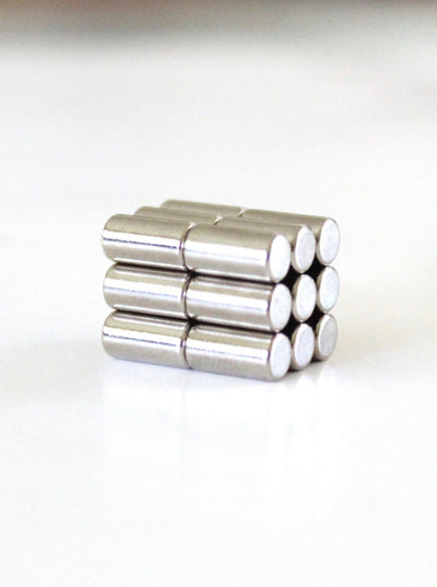 A small stack of Girl Fridays Ultra Strong Neodymium Magnets forms a 3x3 grid on a smooth, white surface. Each magnet is silver with a shiny metallic finish.