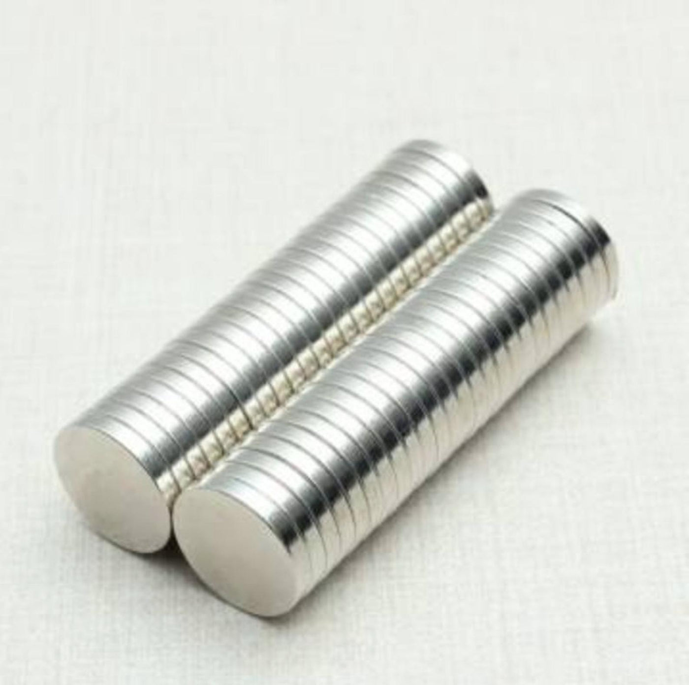 Two stacks of Girl Friday Shops strong neodymium disk magnets, each small and round with a shiny metallic finish and N35 strength, are arranged side by side on a light surface. Their cylindrical shape is clearly visible.