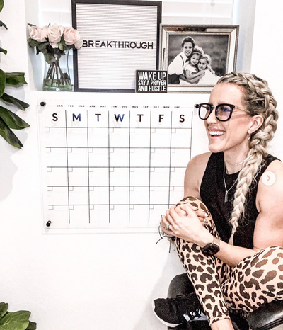 A person with braided hair and glasses is smiling next to a Girl Friday Shop Acrylic Calendar on the wall, which also displays framed photos like one titled BREAKTHROUGH. Theyre wearing patterned pants and sneakers, with a plant nearby.