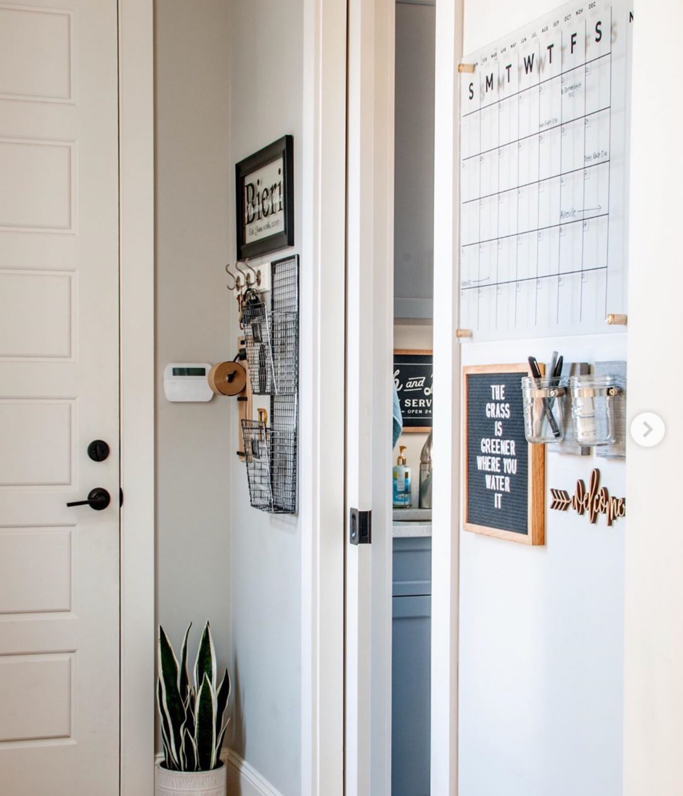 A hallway showcases a Girl Friday Shop Acrylic Calendar - Black text, a wall organizer, and a letter board displaying The grass is greener where you water it. A potted plant sits by the white door with black handles, while wet-erase markers are on the thermostat ledge.