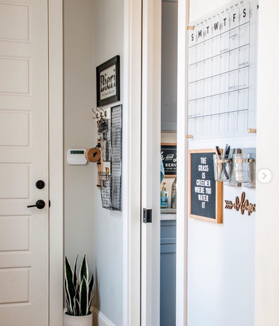 A hallway showcases a Girl Friday Shop Acrylic Calendar - Black text, a wall organizer, and a letter board displaying The grass is greener where you water it. A potted plant sits by the white door with black handles, while wet-erase markers are on the thermostat ledge.
