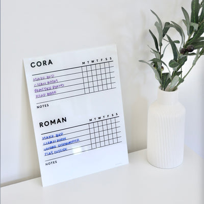 A White Acrylic Chore Chart by Girl Friday Shop sits next to a white vase with green foliage. Customized for two kids, Cora and Roman, it lists tasks like Make Bed and Clean Room in colorful markers, ideal for non-magnetic fridges.
