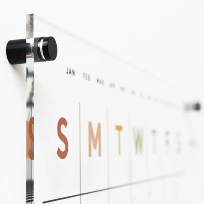 A close-up of the Girl Fridays Acrylic Calendar EARTH TONE RAINBOW features an acrylic wall calendar with months as abbreviations, days in bold colors (S, M, T, W, T, F, S), and sleek black metal pegs. Ideal for wet-erase markers against a softly blurred white background.