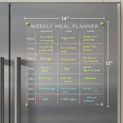 The Girl Friday Shop Clear Acrylic Fridge Meal Planner (14x15) attaches to the fridge, featuring meal options for Monday to Sunday, including breakfast, lunch, and dinner like French toast and chili. Easily update your plan weekly with wet-erase markers for a fresh start.