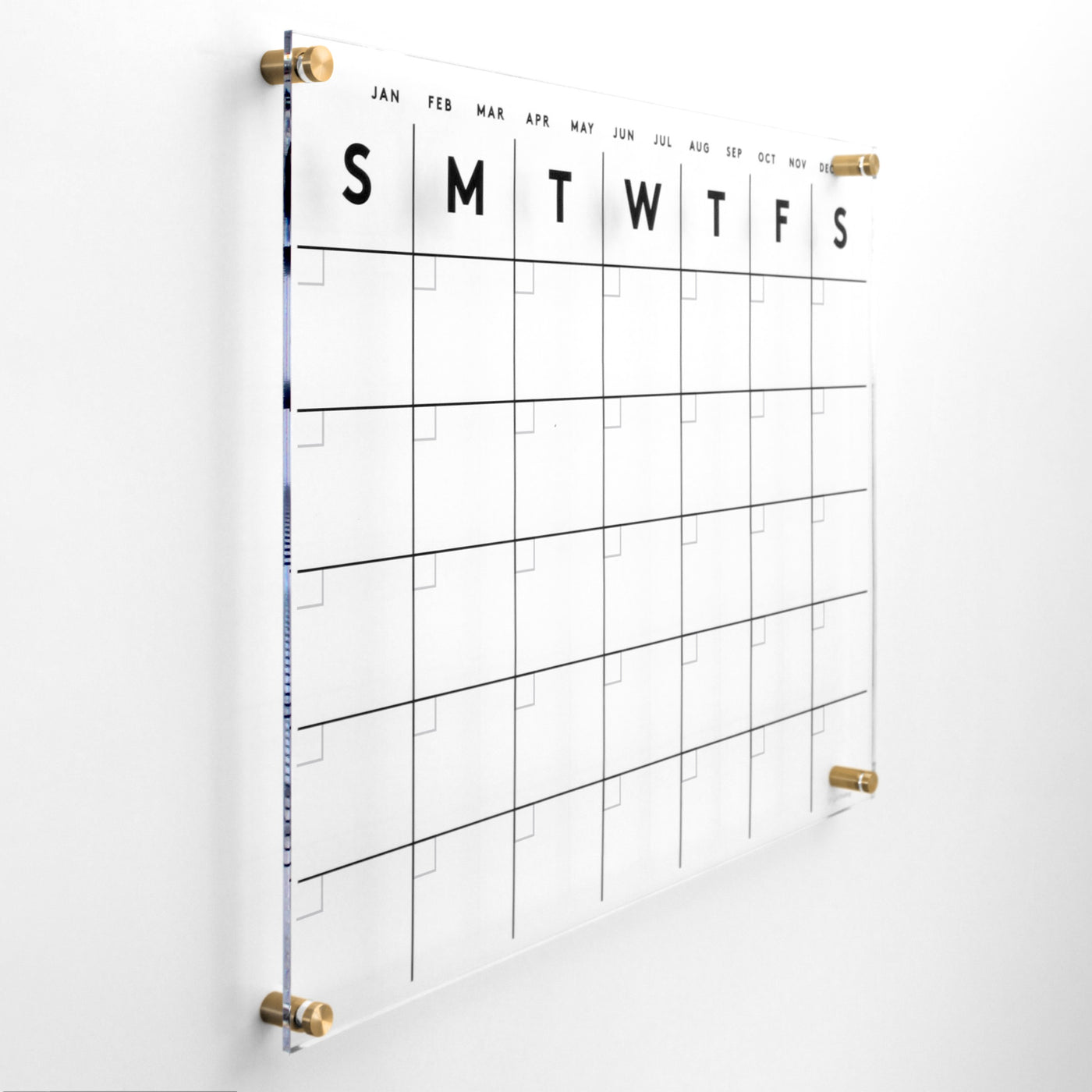The Acrylic Calendar by Girl Friday Shop features a minimalist grid layout displaying days from Sunday to Saturday. Date and event entries use wet-erase markers to prevent ghosting. Months are labeled on top, and its elegantly mounted with gold fasteners.
