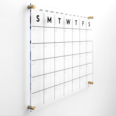 The Acrylic Calendar by Girl Friday Shop features a minimalist grid layout displaying days from Sunday to Saturday. Date and event entries use wet-erase markers to prevent ghosting. Months are labeled on top, and its elegantly mounted with gold fasteners.