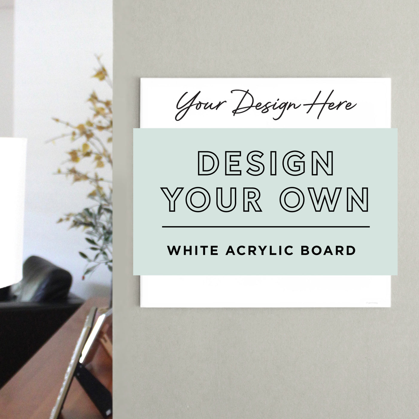 On a gray wall, the White Acrylic Dry Erase Board by Girl Friday Shop showcases Your Design Here and Design Your Own White Acrylic Board in bold black letters. A chair, a lamp, and a plant with yellow flowers are in the background.