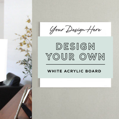 On a gray wall, the White Acrylic Dry Erase Board by Girl Friday Shop showcases Your Design Here and Design Your Own White Acrylic Board in bold black letters. A chair, a lamp, and a plant with yellow flowers are in the background.