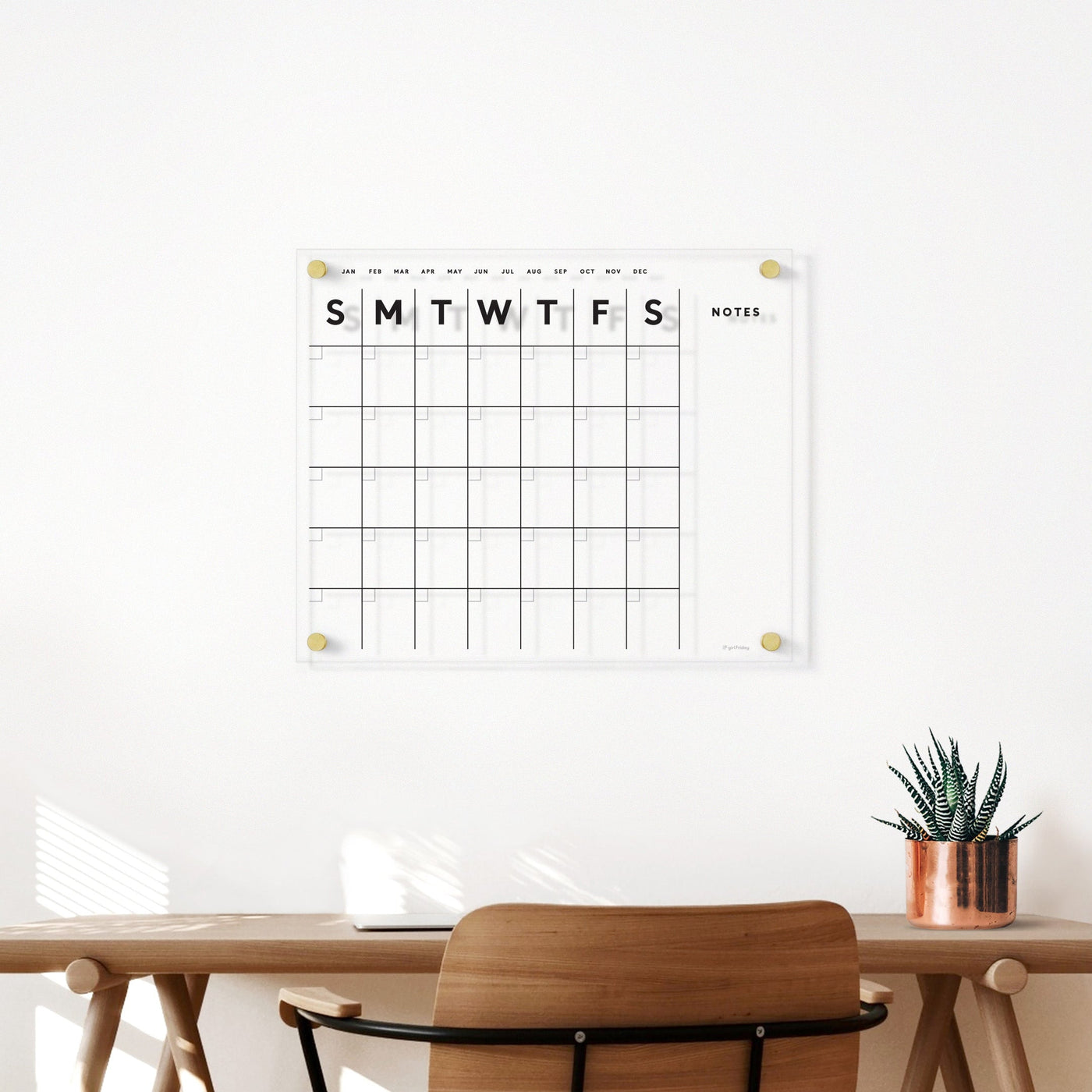 A minimalist office setup with a wooden desk and chair includes a small potted plant. Above, the Girl Friday Shops Clear Acrylic Monthly Wall Calendar with Notes is mounted on the white wall, featuring days of the week and a notes section for fluid planning using wet-erase markers.