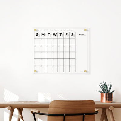 A minimalist office setup with a wooden desk and chair includes a small potted plant. Above, the Girl Friday Shops Clear Acrylic Monthly Wall Calendar with Notes is mounted on the white wall, featuring days of the week and a notes section for fluid planning using wet-erase markers.