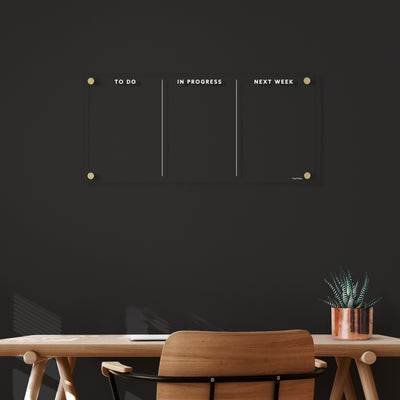 A minimalist office setup includes a wooden desk and black chair. Above, Girl Fridays customizable Acrylic To Do Board features white text in columns labeled To Do, In Progress, and Next Week. A set of wet-erase markers is nearby, while a small potted plant sits at the desks edge.