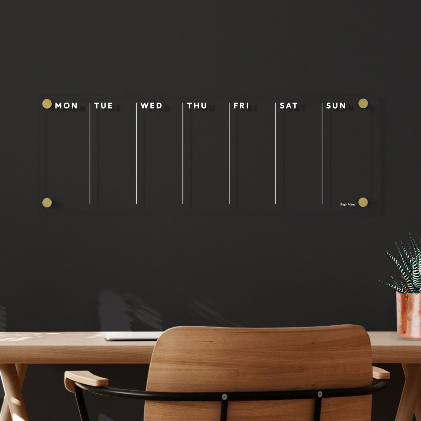 A minimalistic desk setup features the Clear Acrylic Weekly Calendar with white text from Girl Friday Shop, days labeled Monday through Sunday. It includes a potted plant and acrylic organizer, complementing the wooden chair against a dark wall for a clean, modern look.