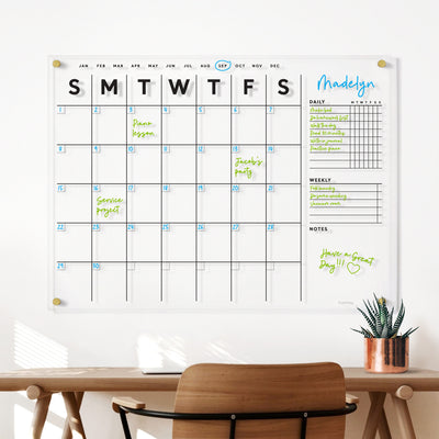 The Clear Acrylic Large Customizable Monthly Wall Calendar with Chore Charts by Girl Friday Shop, designed for families, features notes like soccer party and piano lesson. A wooden desk with a chair and plant sits below, warmed by sunlight—ideal for writing plans with wet-erase markers.