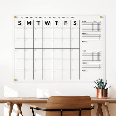 The Clear Acrylic Large Customizable Monthly Wall Calendar with Chore Charts from Girl Friday Shop enhances organization on a white wall above a wooden desk, serving as a stylish family planner. Paired with wet-erase markers, the setup includes a copper-potted plant and a wooden chair in front.