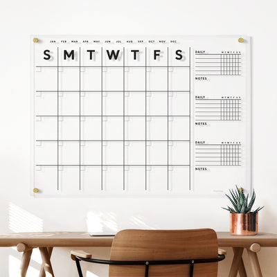The Clear Acrylic Large Customizable Monthly Wall Calendar with Chore Charts by Girl Friday Shop, featuring columns for days and rows for weeks, is mounted above a wooden desk. Enhanced by a sunlight-bathed potted plant, it’s perfect for families and works seamlessly with wet-erase markers.