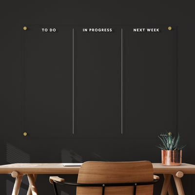 A minimalist workspace showcases a wooden desk and chair. An Acrylic To Do Board by Girl Friday, divided into To Do, In Progress, and Next Week columns in white text, adorns the wall, ideal for wet-erase markers. A small potted plant on the desk adds a touch of greenery.