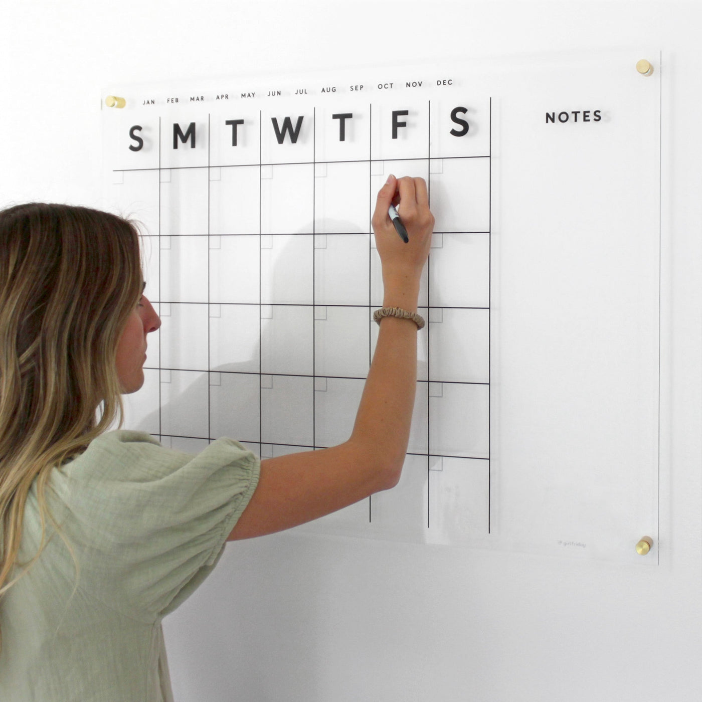 Wearing a green shirt and wristband, an individual with long hair efficiently organizes their familys schedule on the Girl Friday Shops Clear Acrylic Monthly Wall Calendar with Notes. They use vibrant wet-erase markers on the wall-mounted planner, featuring weekly headers, empty grids below, and a Notes section on the right.