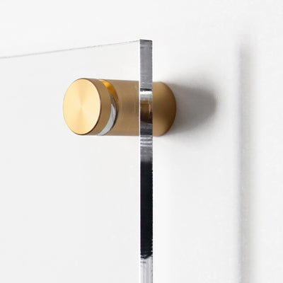 A close-up shows a Clear Acrylic Large Customizable Monthly Wall Calendar with Chore Charts by Girl Friday Shop, mounted on a wall with round, brass-colored hardware brackets. The minimalist design contrasts the transparent surface with the metallic finish while enhancing functionality.