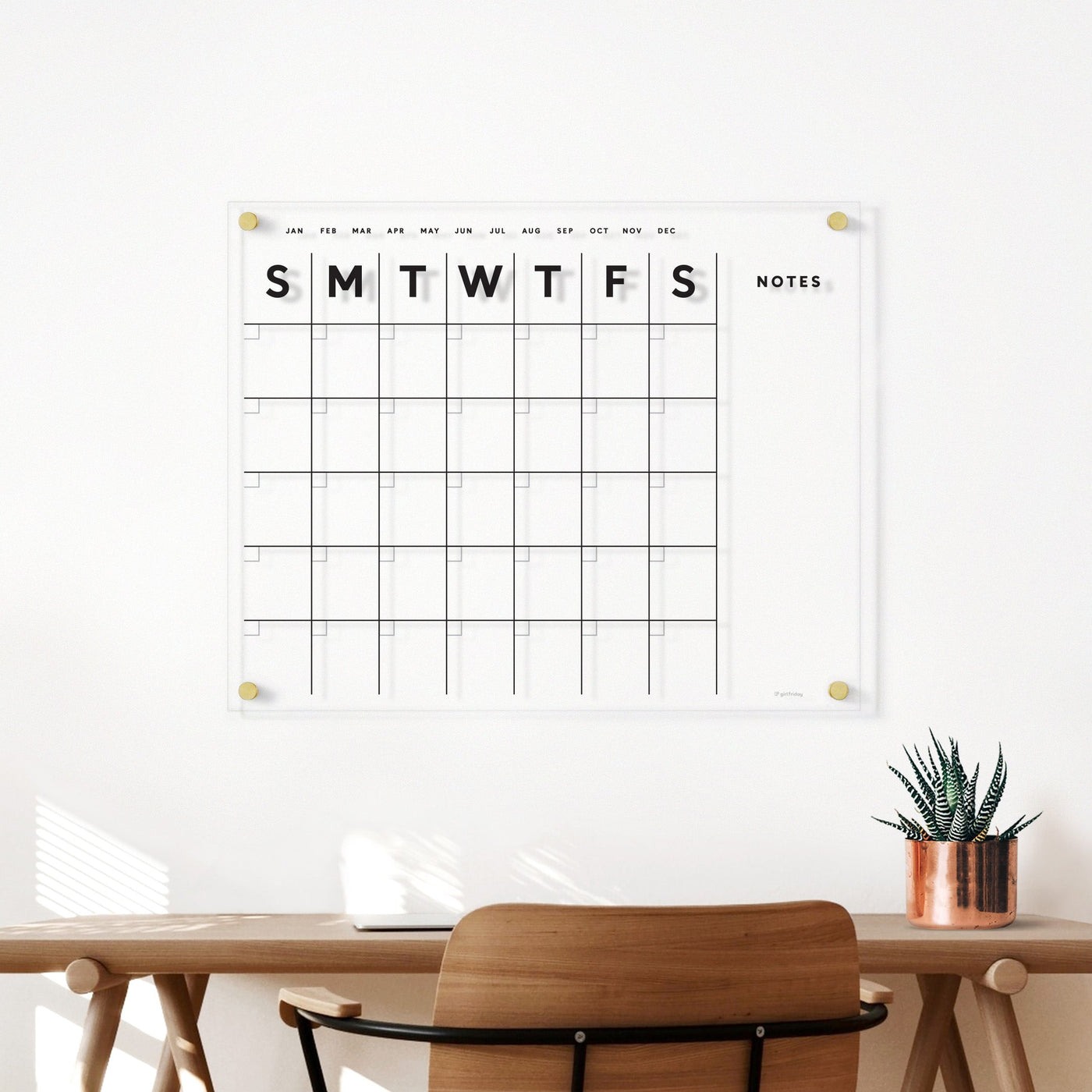 The Girl Friday Shops Clear Acrylic Monthly Wall Calendar with Notes, featuring bold weekly grids, is mounted above a wooden desk and brown chair. Wet-erase markers and a potted plant are neatly placed on the desk, making it ideal as a family planner.