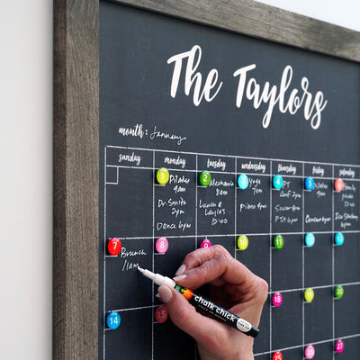 Using a CalendarGirlCompany 24 x 36 Large Vertical Chalkboard, someone writes on The Taylors black chalkboard calendar with chalk ink markers. The January schedule, adorned with colorful sticky notes and events like an 11 am brunch, blends organization and creativity seamlessly.