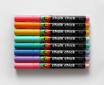 Eight Chalk Ink Vibrant Markers from Girl Friday, featuring vibrant colors like pink, red, and teal, rest on a white background. Colorful caps add flair to a stainless steel cup holder. Each label displays Chalk Chick, making them functional and stylish.